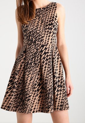 Mouwloze beige jurk met uitlopende rok en een abstract zwart pied-de-poule patroon, gedragen door een vrouw met lang bruin haar.