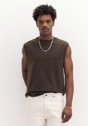 Göndör hajú férfi sötétbarna ujjatlan ingben, fehér nadrágban, ezüst láncnyaklánccal és karkötővel, fehér háttér előtt áll.