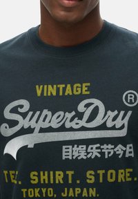 Zwart katoenen T-shirt met ronde hals en een groot vintage-stijl "SuperDry" logo in zilver, geaccentueerd met gele tekst en Aziatische karakters.