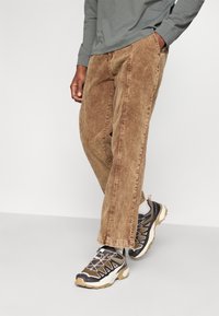 Brązowe spodnie kordowe o szerokim kroju, z widocznym przeszyciem. Połączone z czarnymi i beżowymi trampkami z fakturowanymi akcentami.