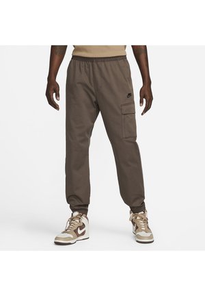 Man in een bruine Nike cargobroek en beige, bruine en witte Nike sneakers, staand tegen een witte achtergrond.
