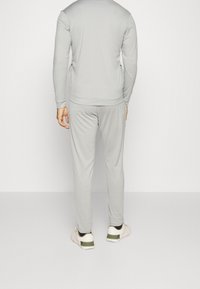 Homme debout, tourné de dos, portant un survêtement gris clair avec des baskets blanches sur un fond clair uni.