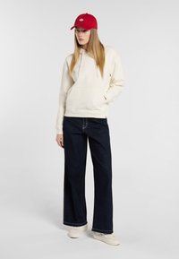 Felpa in cotone crema con tasca frontale; abbinata a jeans a gamba larga blu scuro e sneakers crema; cappellino rosso con dettaglio logo.