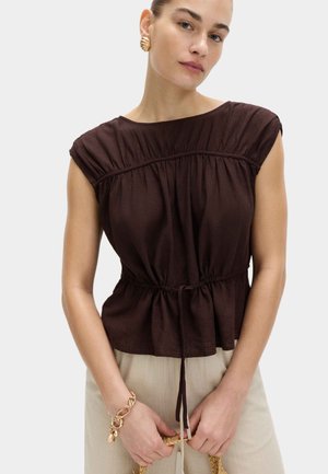 Frau trägt ein dunkelbraunes, gerafftes ärmelloses Oberteil, beige Hose, ein goldenes Kettenarmband und goldene Creolen, hält eine Tasche mit geflochtenem Griff.