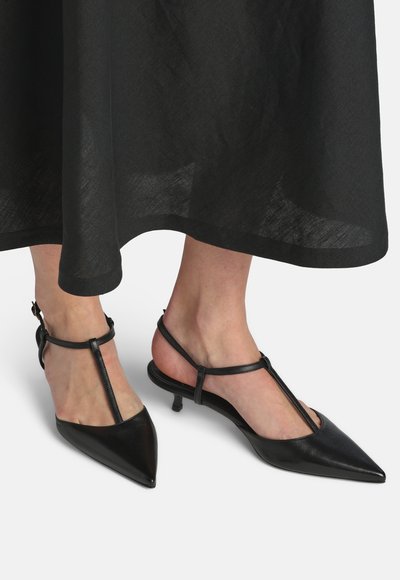 Escarpins à bout pointu en cuir noir avec bride en T, portés avec une jupe noire fluide, dévoilant les chevilles et le bas des jambes.