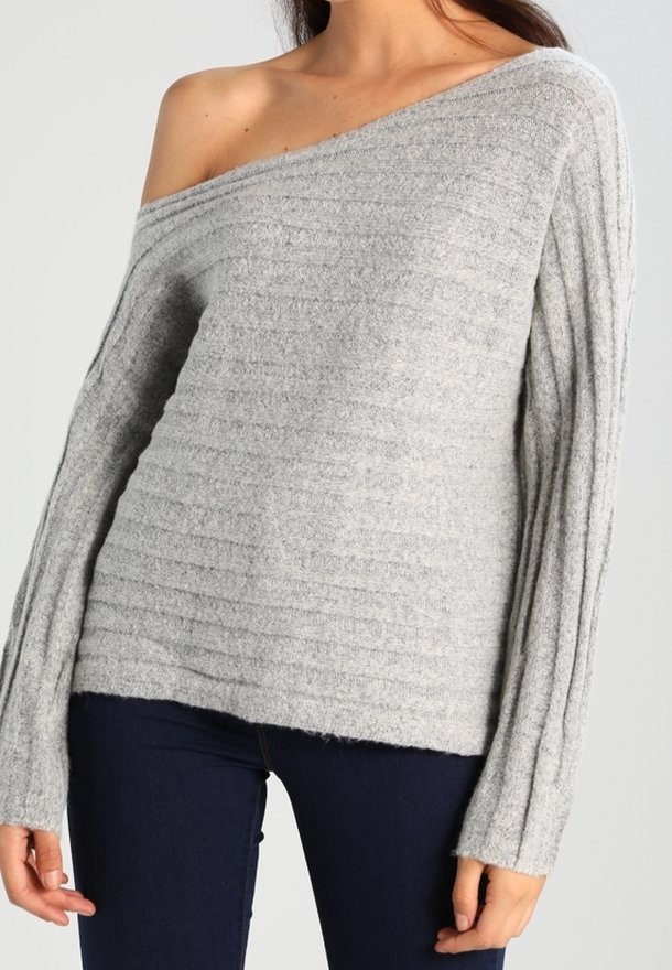 Femme portant un pull gris clair à côtes, en maille, à épaules dénudées, associé à un pantalon ajusté bleu foncé, sur un fond uni.