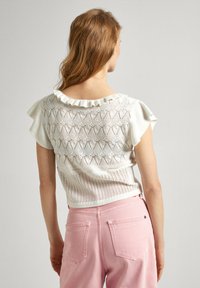 Haut en tricot blanc avec un décolleté à volants et des manches courtes, doté d'un motif ajouré complexe, associé à un pantalon taille haute rose pastel.