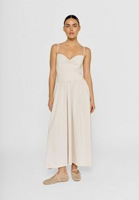 Beige midi-kjole med tynde stropper, samlet bustdetalje, flagrende skørt og blød tekstur, parret med lyse beige slip-on sko.
