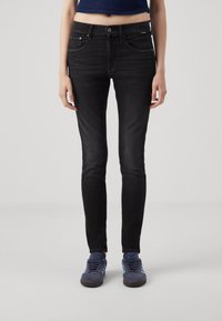 Jeans negros ajustados de denim, con cierre delantero de botón, trabillas para cinturón y cinco bolsillos. Llevados con zapatillas azules.