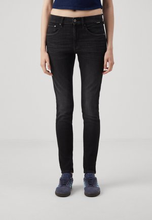 Jeans negros ajustados de denim, con cierre delantero de botón, trabillas para cinturón y cinco bolsillos. Llevados con zapatillas azules.