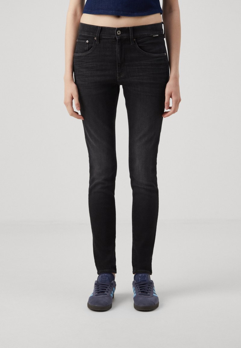 Jeans negros ajustados de denim, con cierre delantero de botón, trabillas para cinturón y cinco bolsillos. Llevados con zapatillas azules.