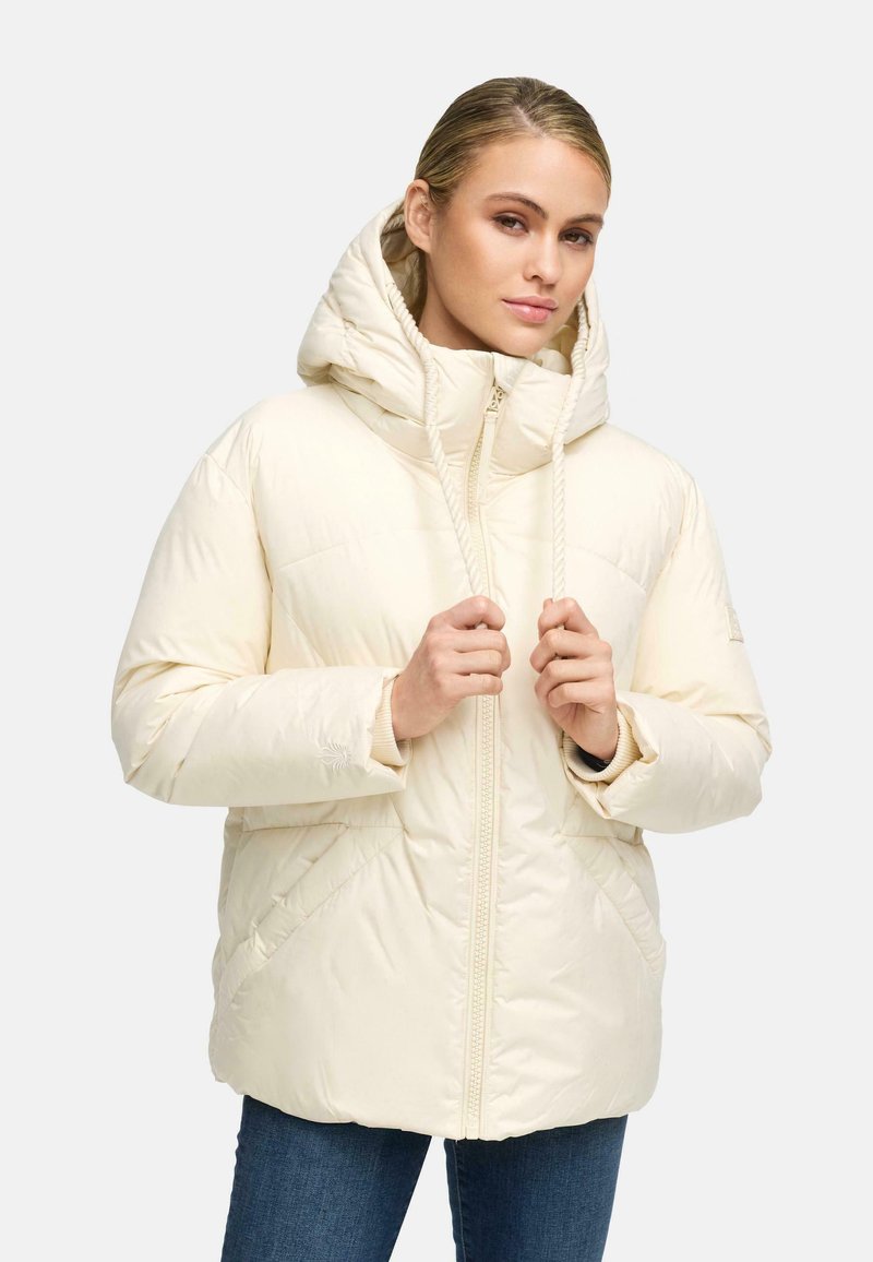 Woman wearing a cream-colored puffer jacket with hood, holding drawstrings, paired with blue jeans on a plain light background.