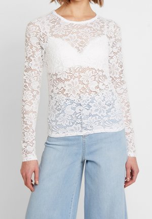 Biały, długi rękaw z kwiatowej koronki noszony na białym biustonoszu typu bralette, zestawiony z jasnoniebieskimi, wysokim stanem i szerokimi nogawkami jeansami.