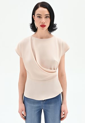 Donna con capelli neri fino alle spalle che indossa un top beige drappeggiato, jeans blu, rossetto rosso e un singolo orecchino lungo dorato.