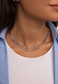 Srebrny metalowy naszyjnik z wydłużonymi owalnymi ogniwami, o gładkim wykończeniu. Noszony na szyi, w połączeniu z jasnoniebieską koszulą.