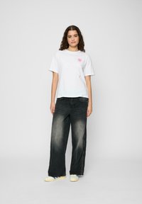 Tricou alb din bumbac cu grafică florală roz, asortat cu blugi negri largi și adidași galbeni cu accente albastre.