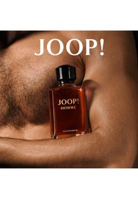 Brun glassflaske med svart lokk, merket "JOOP! HOMME," som hviler på solbrun hud. Klar bakgrunn med teksten "JOOP!" over flasken.