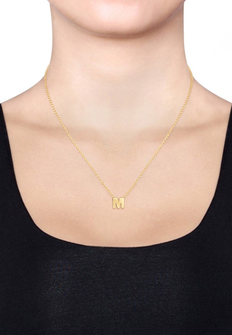 Collana in metallo dorato con un pendente a "m" minuscolo, caratterizzata da una delicata catena e da una finitura lucida, indossata sulla pelle nuda.