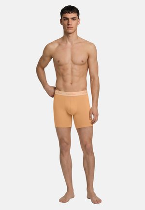 Młody mężczyzna o krótkich ciemnych włosach, ubrany w dopasowane, brzoskwiniowe bokserki z logo Calvin Klein na pasie, stojący boso.