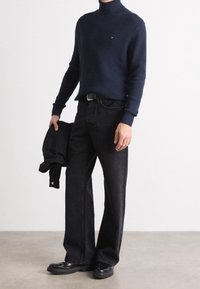 Homme portant un pull à col roulé bleu marine, jean noir à jambes larges, chaussures noires, tenant une veste noire pliée contre un mur blanc.