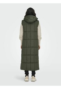 Person wearing a long, hooded, dark green quilted vest over a cream sweater, black pants, and beige sneakers, standing facing away.