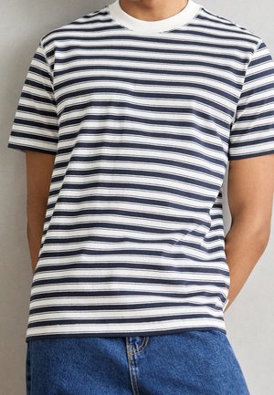 Person trägt ein weißes und dunkelblaues horizontal gestreiftes Kurzarmshirt mit blauen Jeans, steht vor einem einfarbigen Hintergrund.