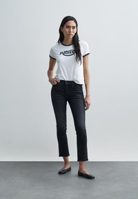 T-shirt bianca con collo a contrasto nero e grafica "MOTHER", abbinata a jeans scuri in denim e ballerine nere, in piedi davanti a uno sfondo semplice.