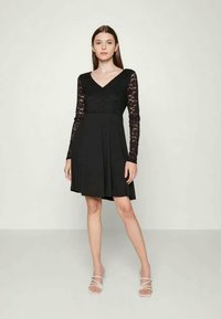 Jovem mulher de pé, vestindo um vestido preto com mangas longas de renda e decote em V, calçando sandálias brancas de tiras, em um fundo claro.