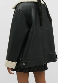 Chaqueta de cuero sintético negra con un suave cuello y puños de borrego crema, que presenta un corte holgado, costuras verticales y un detalle de herrajes circulares.