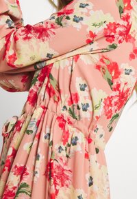 Detailní záběr na broskvové šaty s červenými, žlutými a modrými květinovými vzory a nařaseným pasem, které má na sobě osoba.