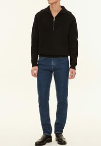 Svart ribbad stickad tröja med halvdolkt krage, kombinerad med blå denimjeans och svarta loafers. Enkel, skräddarsydd passform; avslappnad look.