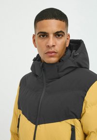 Schwarz-gelbe isolierte Jacke mit hohem Kragen, Frontreißverschluss und Reißverschlusstaschen; Rautensteppmuster im gelben Bereich.