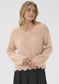 Maglione rosa chiaro, lavorato a maglia, con scollatura a V, maniche lunghe e orlo a onde. Texture morbida con vestibilità ampia. Indossato sopra una gonna scura.