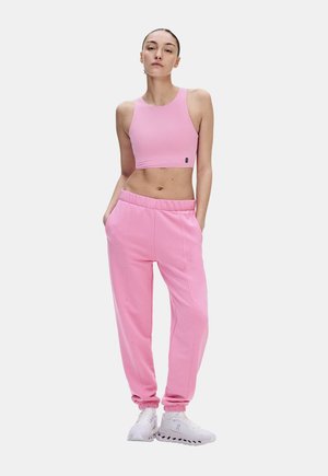 Femme debout, les mains dans les poches, portant un crop top rose, un pantalon de jogging rose et des baskets blanches sur un fond uni.