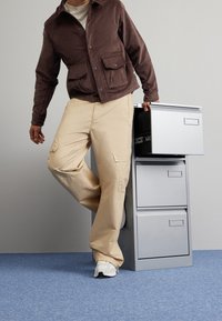 Metallskåp i silver med tre lådor. En person i brun jacka och beiga byxor interagerar med skåpet. Blått matt bakgrund.