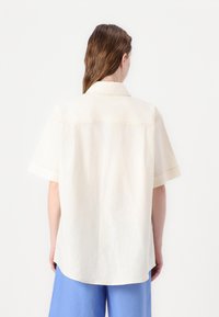 Camicia a maniche corte senza colletto in tessuto beige chiaro. Presenta una vestibilità dritta con un dettaglio di cucitura sulla schiena e aperture laterali.