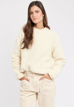 Pull en tricot torsadé couleur crème avec un col montant et une coupe décontractée, texturé avec un motif en diamant, porté avec un pantalon beige.