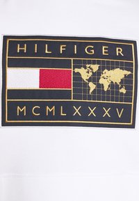 Tessuto bianco con un logo rettangolare blu navy. Ricamo dorato che include "HILFIGER," un blocco rosso e bianco, e una mappa del mondo con "MCMLXXXV."