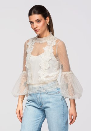 Femme aux cheveux bruns en queue de cheval portant un chemisier en dentelle blanche transparente avec des manches bouffantes et un jean bleu clair sur un fond uni.