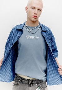 Cămașă deschisă cu carouri albastre purtată peste un tricou alb cu inscripția "SWOM". Blugi gri și coliere suprapuse cu diverse pandative.