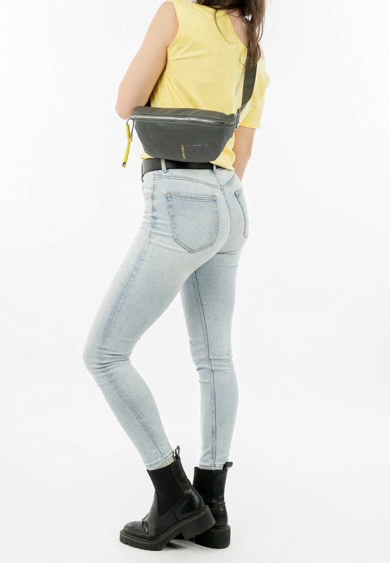 Personne portant un jean bleu clair, des bottines noires et un top jaune sans manches, portant un sac bandoulière noir avec une fermeture éclair jaune.