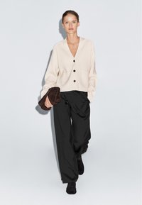 Beige gebreide cardigan met een V-hals, voorzien van vier donkere knopen en twee voorkwaliteiten, gecombineerd met losse zwarte broeken en een bruine clutch.