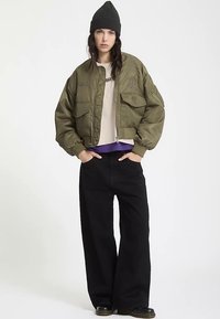 Giacca bomber corta verde oliva con dettagli ricamati, abbinata a una camicia color crema e pantaloni neri larghi. Il berretto nero completa il look.