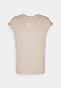 Camiseta beige de algodón con cuello redondo y mangas cortas. Presenta un logo blanco de "ALPHA INDUSTRIES" en el pecho.