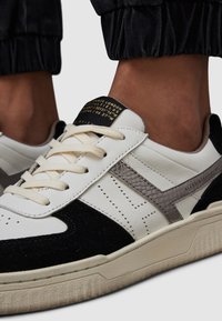 Bílé kožené tenisky s černými semišovými akcenty, šedým strukturovaným bočním pruhem, šněrovacím designem a polstrovaným jazykem s zlatým brandingem.