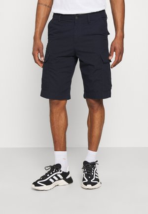 Homme portant un short cargo bleu marine, des chaussettes blanches et des baskets noir et blanc, debout devant un fond clair uni.