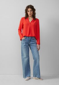 Rode blouse met lange mouwen en v-hals, gecombineerd met lichtblauwe wijde spijkerbroek. Model draagt lichtgekleurde instappers. Eenvoudig ontwerp, casual stijl.