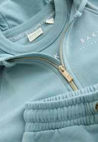 Svetlomodrá mikina Ted Baker so zlatým zipsom, viditeľnou značkou a elastickým pásom nohavíc alebo šortiek v rovnakej farbe.