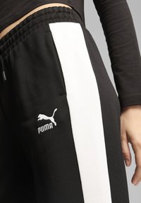 Čierne tepláky so bielym bočným prúžkom, vyrobené z hladkého materiálu, s vyšitým logom Puma na stehne. Viditeľný elastický pás.