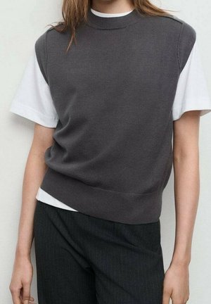 Persoon draagt een donkergrijze mouwloze gebreide vest over een wit T-shirt met korte mouwen en donkere broek met krijtstrepen, tegen een effen achtergrond.